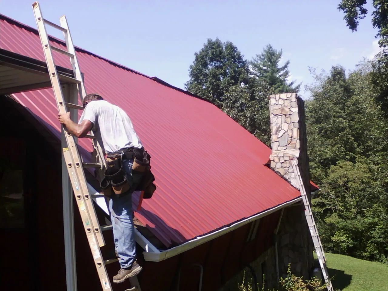 Roofer on a ladder beside a red metal roof and stone chimney — Soffit & Fascia Repair by Roofing Brothers in Elkwood